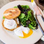 Two poached eggs on a white plate with toasted English muffin and some salad greens, knife and fork.