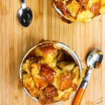 Two ramekins of bread pudding with two spoons on a bamboo surface.