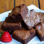 A stack of brownies on a whit plate with a strawberry on the side.