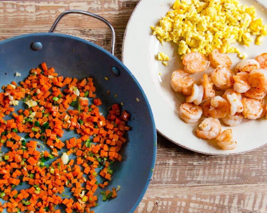 A pan with sautéed carrots next to a plate with cooked shrimp and egg.