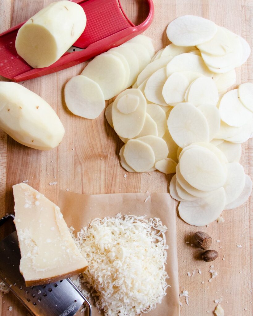 Ingredients for potato gratin on a wooden cutting board - potatoes, parmesan cheese, nutmeg - and a mandolin.