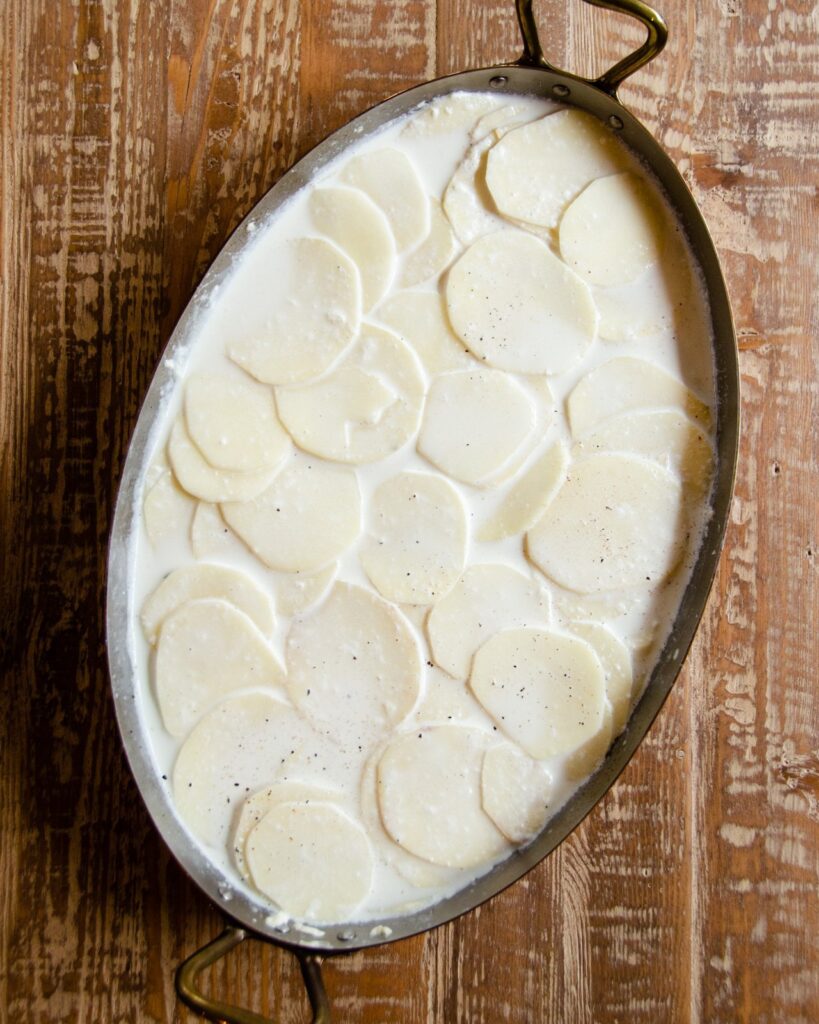 Potatoes and half-and-half in a copper oval gratin pan, uncooked.