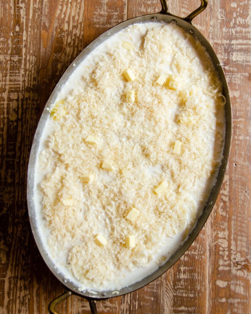 Potatoes and half-and-half in a copper oval gratin pan, uncooked with Parmesan cheese on top.