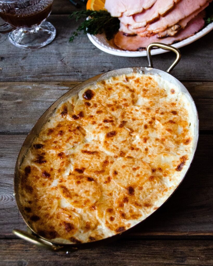 Potato gratin in a copper oval gratin pan on a wooden table with ham in background.