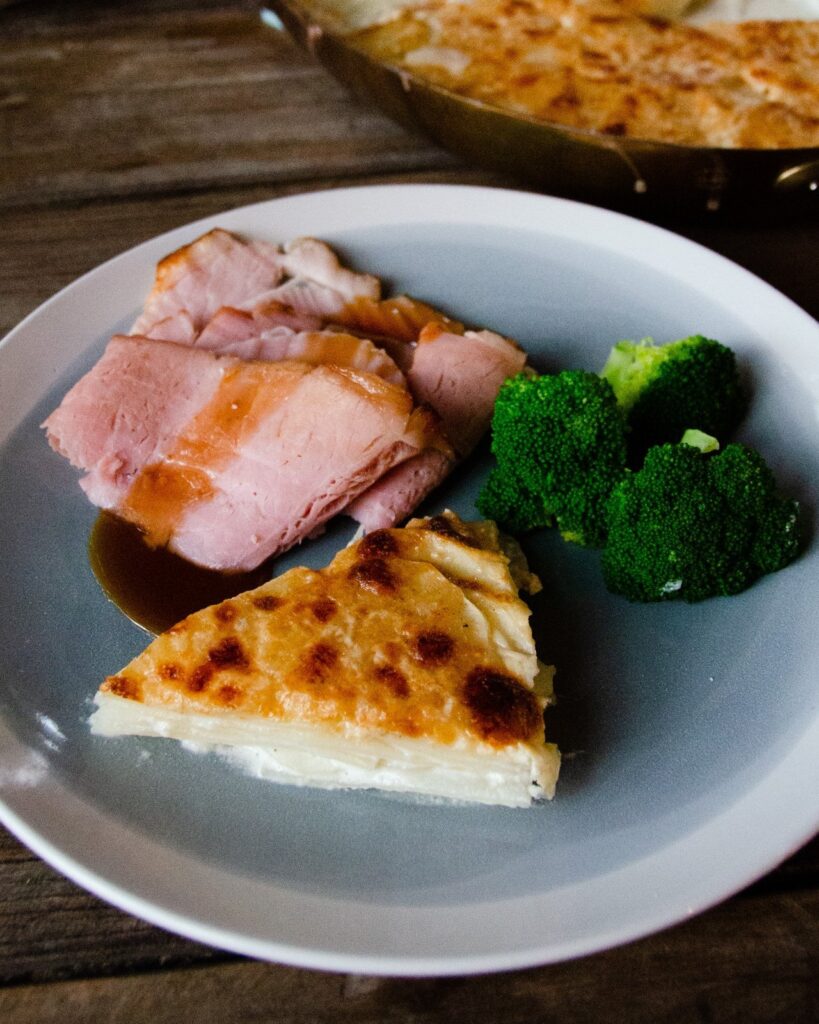 A blue dinner plate with a wedge of potato gratin, ham and broccoli.