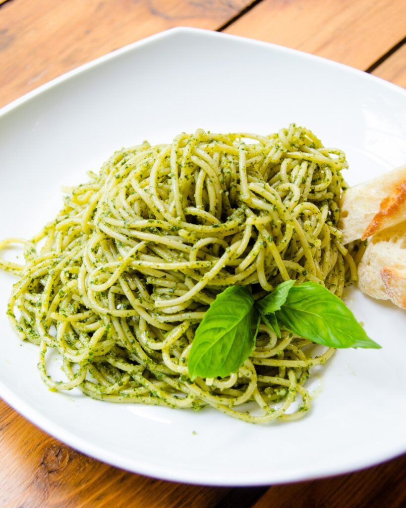 Spaghetti with basil pesto on a white plate with bread and two leaves of basil.