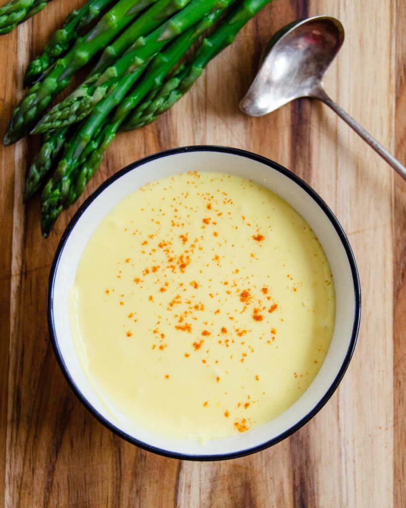 A bowl of hollandaise sauce with asparagus and a ladle near by.