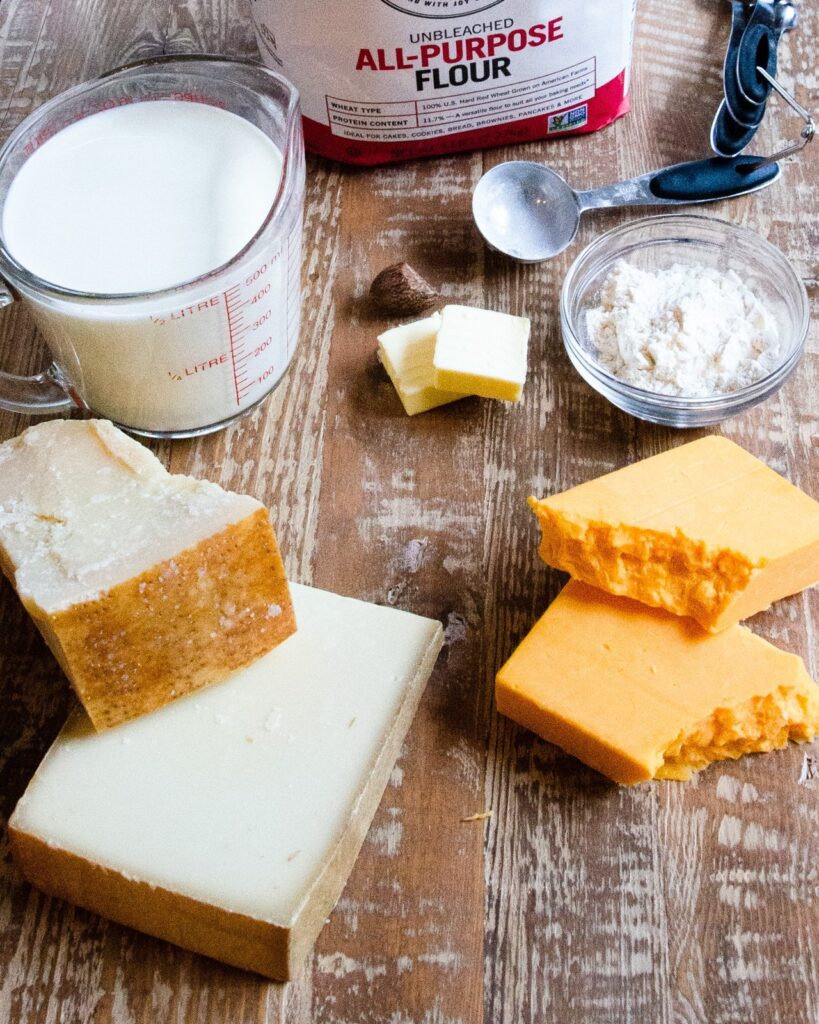Ingredients for cheese sauce on a wooden counter - Parmesan, Gruyère, Cheddar, milk, butter, flour and nutmeg.