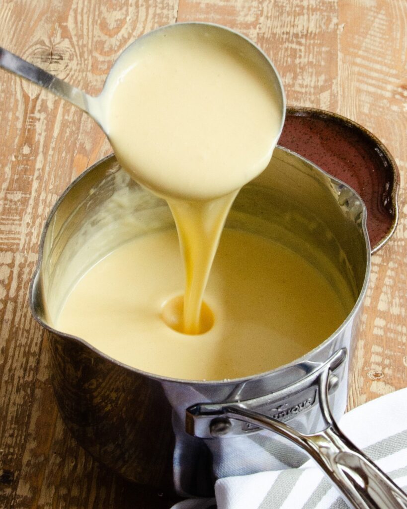 A ladle pouring cheese sauce into a saucepan.