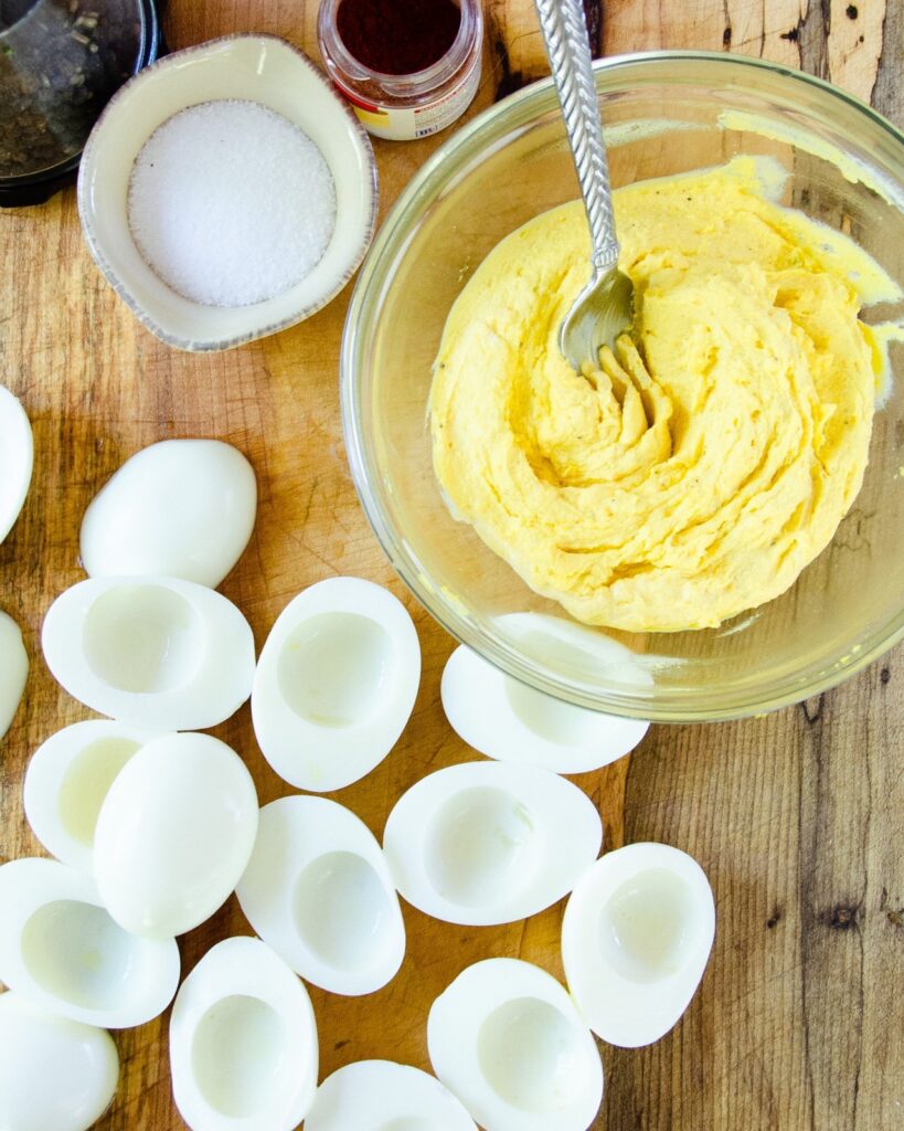 Hard boiled egg whites on a wooden cutting board with a bowl of deviled egg yolk filling.