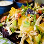 Nachos with chicken, avocado, scallions and salsa on a black platter.