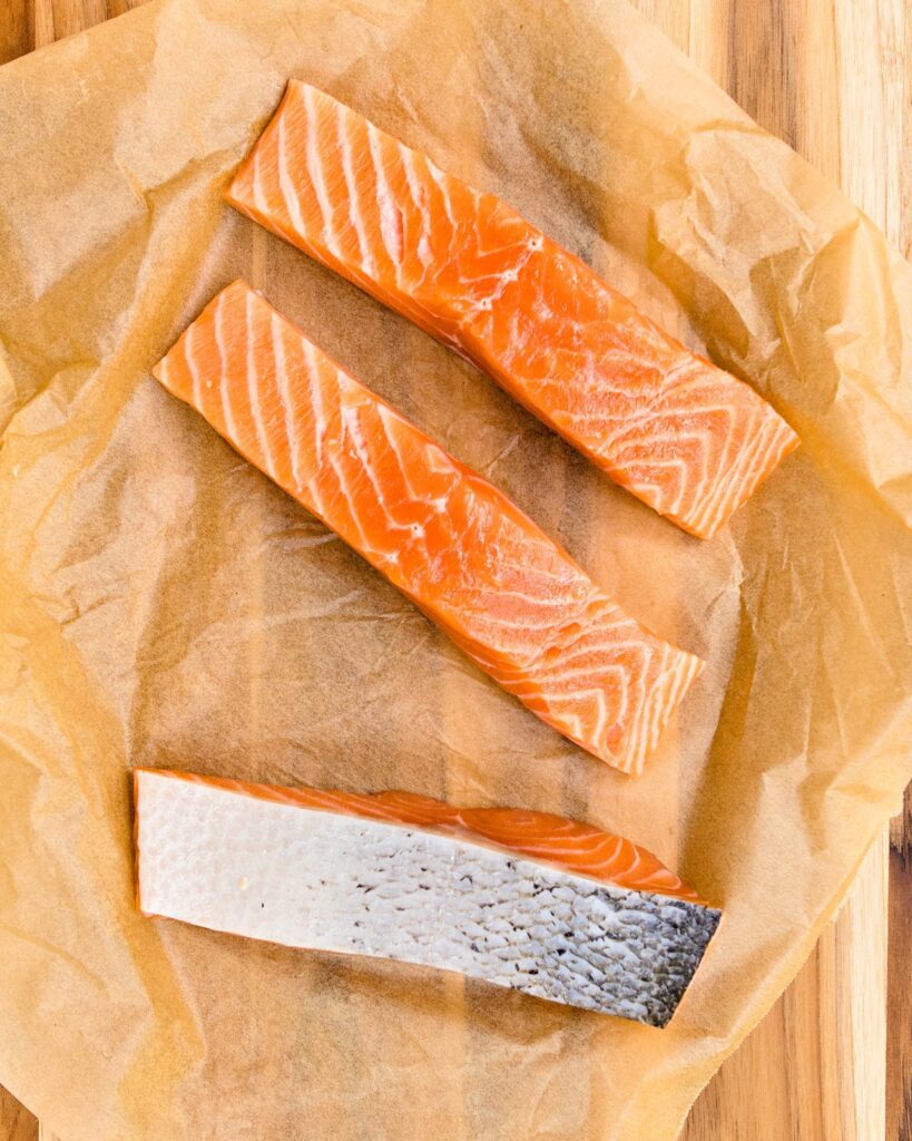 Three raw fillets of salmon on brown parchment on a wooden cutting board.