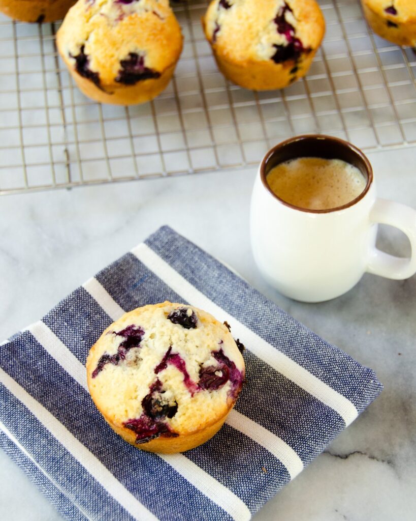 A blueberry muffin on a blue and white striped napkin and an espresso.