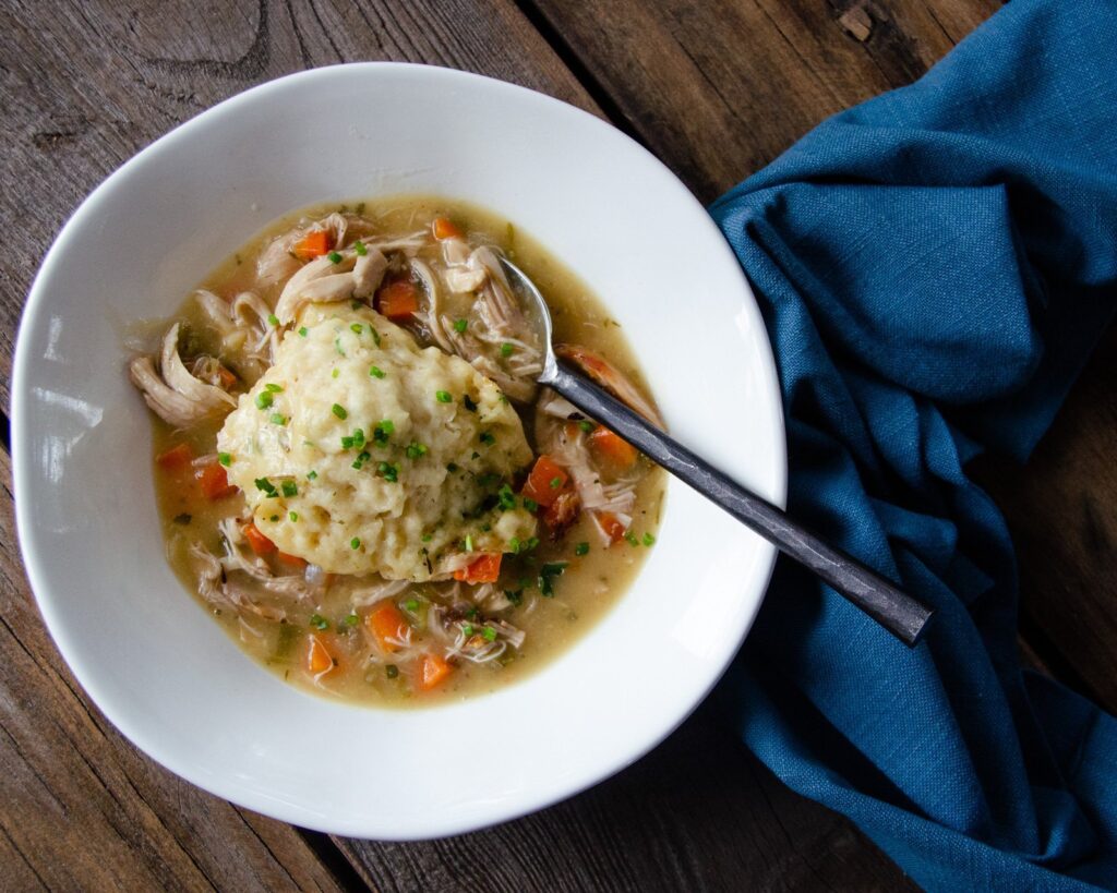 A white bowl with chicken and dumplings in it, along with a spoon and a blue napkin.