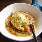 A white bowl with chicken and dumplings and a spoon ion a wooden table.