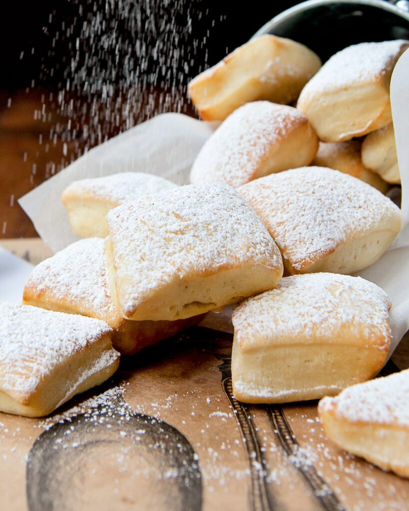 A bucket of air-fried beignets tipped over onto a table with powdered sugar dusting down from above.