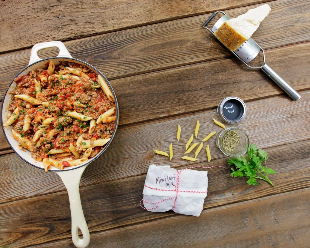 A pot of one pot bolognese with a package of meatloaf mix, some dried penne, some parsley, fennel seed and Parmesan cheese on a wooden table.