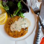 One crab cake with sherry sauce on a white plate with lemon wedges and mixed greens.