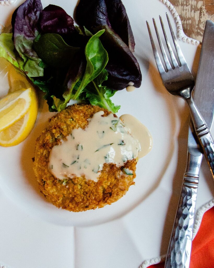 One crab cake with sherry sauce on a white plate with lemon wedges and mixed greens.