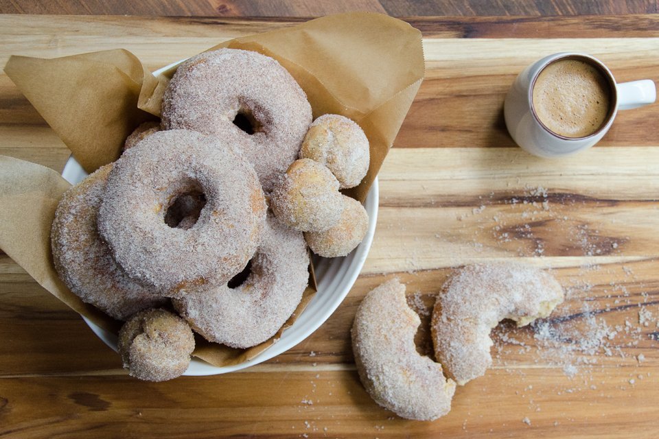 Bowl of air-fried apple cider doughnuts with one broken open on a cutting board and a cup of coffee on the side.
