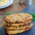 Loaded cauliflower hash browns stacked on a slate tray with eggs on a plate in the background.