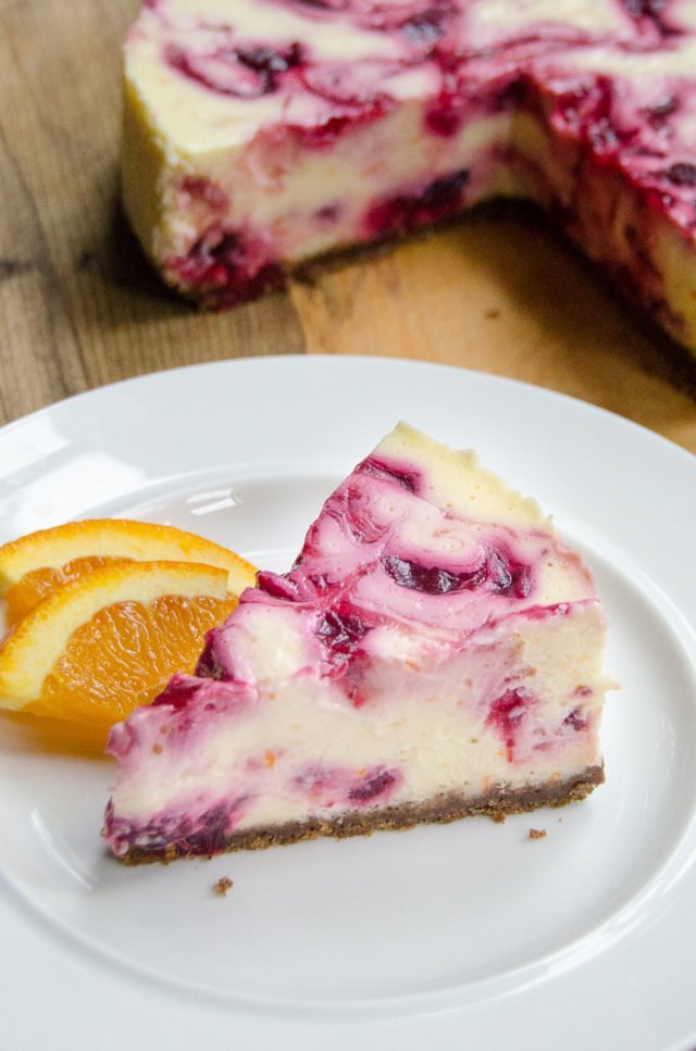 A wedge of Orange Cranberry Cheesecake on a white plate with the whole cake in the background.