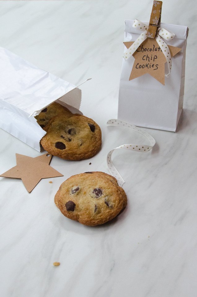 Chocolate chip cookies in a white paper bag on a marble counter with some ribbons and gift tags.
