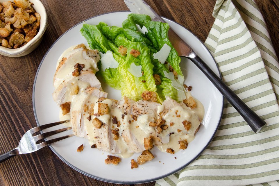 Cream Caesar Chicken chicken breast sliced on a white plate with sauce and bread crumbles on top and romaine lettuce on the side.