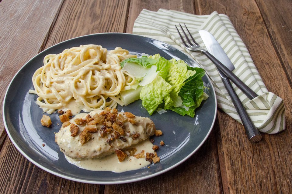 Whole Creamy Caesar Chicken chicken breast on a grey plate with fettuccine and romaine lettuce on the side.
