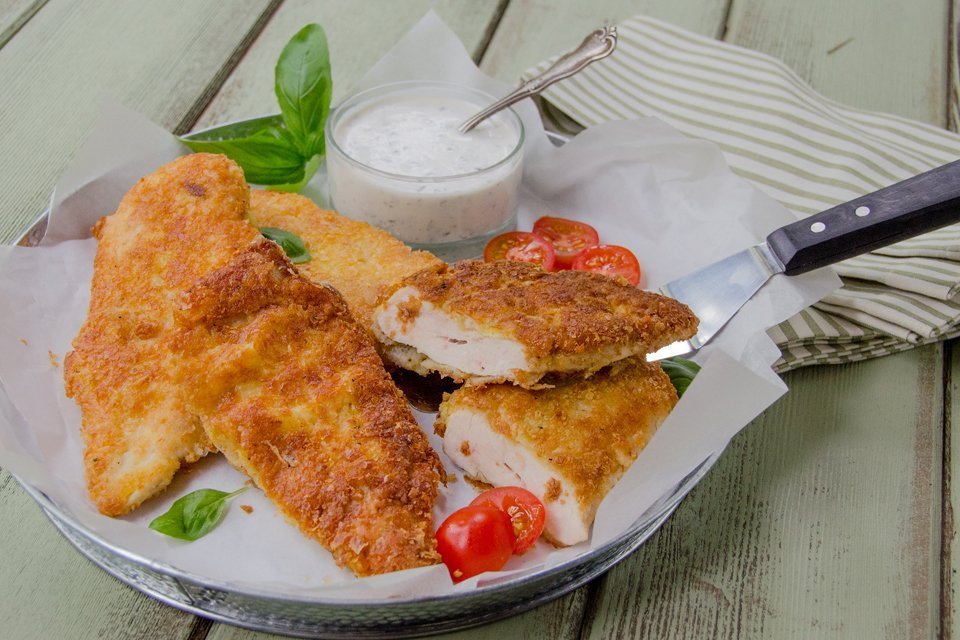 Gluten-free breaded chicken cutlets on a white plate with tomatoes and basil and a basil buttermilk dressing on the side.