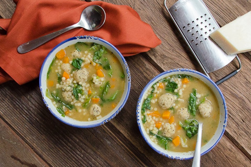 Two bowls of Italian Wedding Soup on a wooden table with an orange napkin and some Parmesan cheese and microplane.