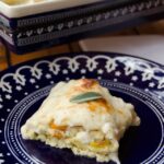 A piece of roasted squash lasagna on a blue plate with a baker in the background.