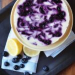 Lemon Blueberry Cheesecake on a cake stand with lemons and blueberries next to it.