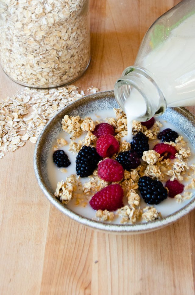 Homemade oat milk being poured over a bowl of granola with berries.