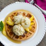 Peach ice cream on waffles on a white plate with a pink napkin and fork.