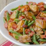 Potatoes and green beans with bacon in a white bowl on a burlap runner.