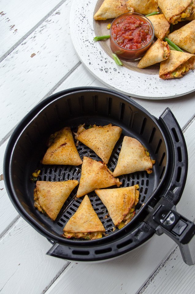 Air-fried chicken stuffed tortillas in an air fryer basket with a plate of finished tortillas nearby.