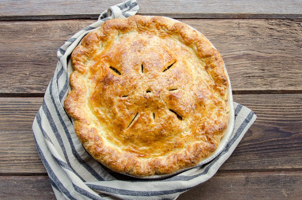 A cooked tourtière on a wooden table with a napkin around the pie dish.