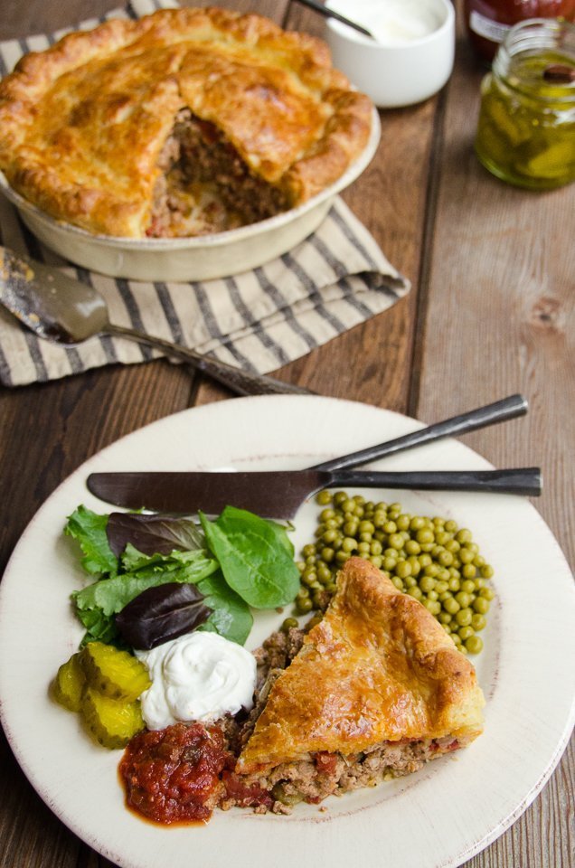 A white plate with a piece of tourtière, some Le Sueur peas and condiments.