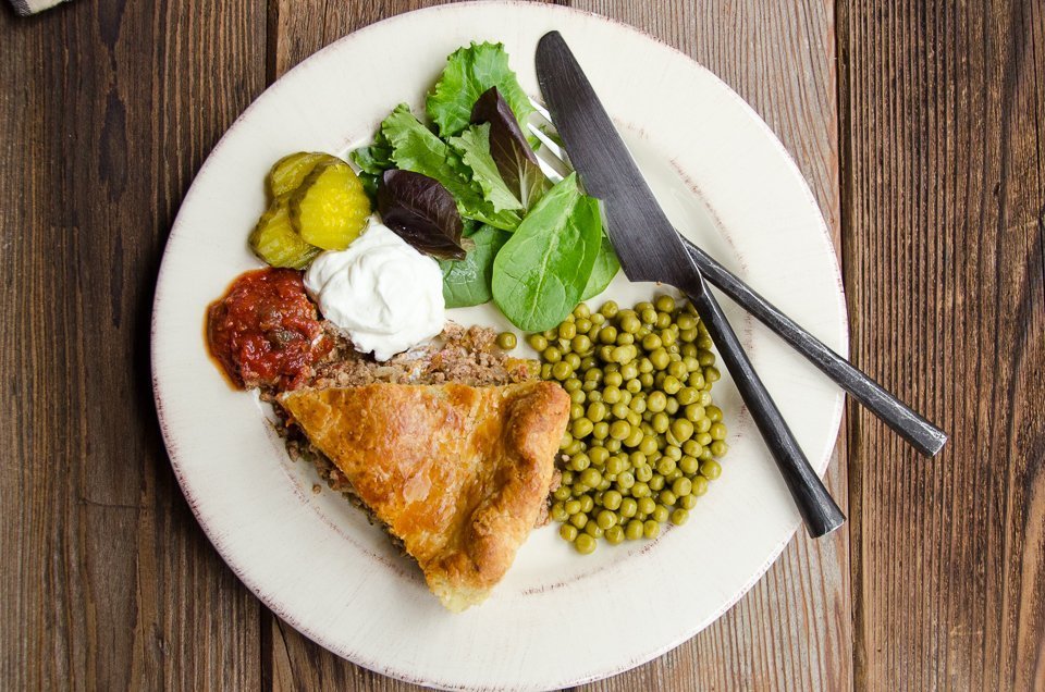 A white plate with a piece of tourtière, some Le Sueur peas and condiments.