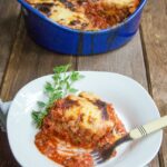 A portion of Vegetarian Moussaka on a white plate with the whole casserole behind.