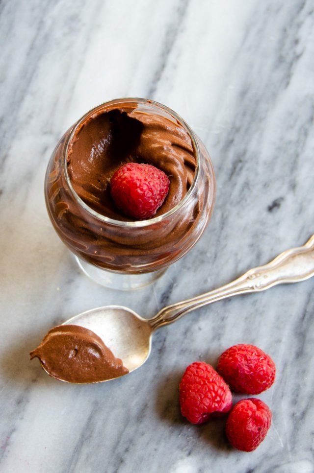 One glass dish with Hervé This' chocolate mousse on a marble countertop with raspberries and a silver spoon.