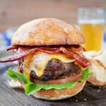 Bourbon bacon burger pn a wooden plate with a beer in the background
