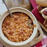 A pottery crock filled with Baked Beans and hot dogs near by.