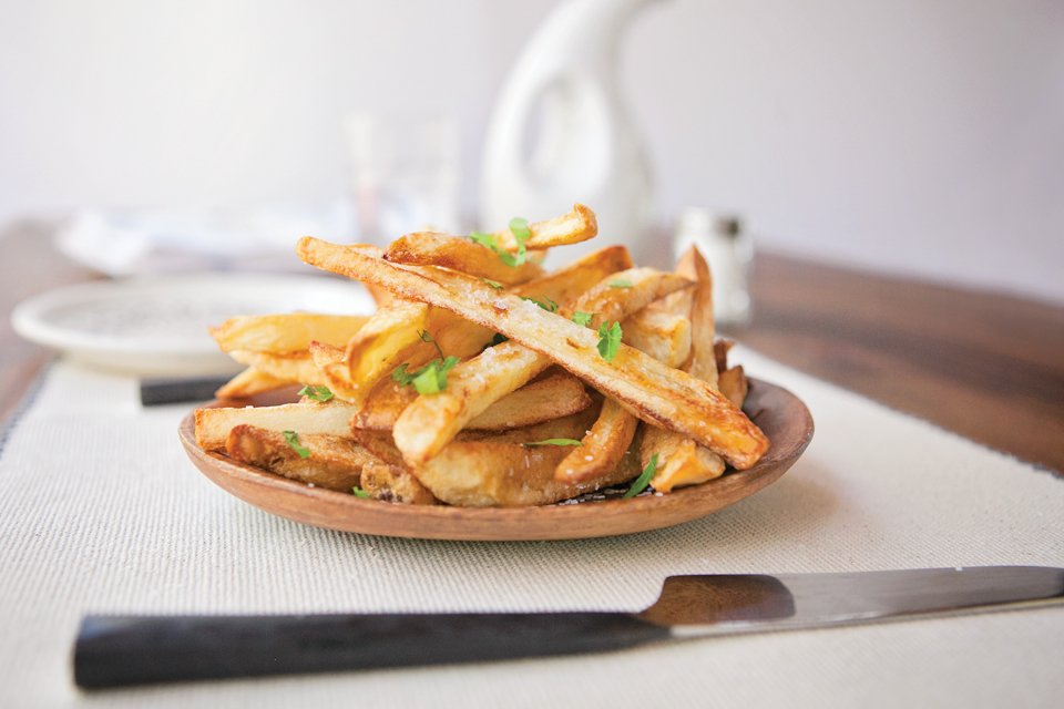 Homemade air-fried French fries on a wooden dish on a white placemat with salt and oil in the background.