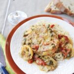 Pork Chops with artichokes and sun dried tomatoes over angle hair pasta on a white plate with place setting and a glass of wine.