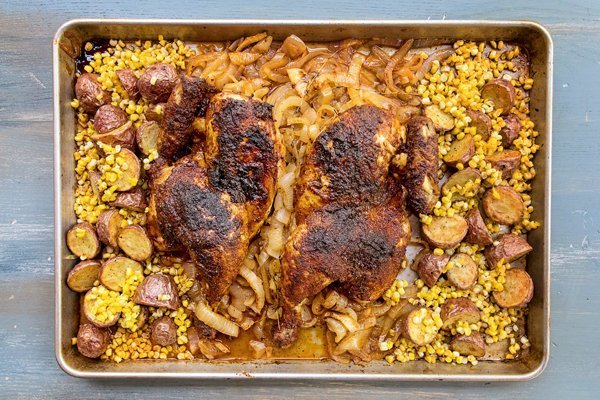 Two summer ale chicken halves on a sheet pan surrounded by onions, corn and potatoes.