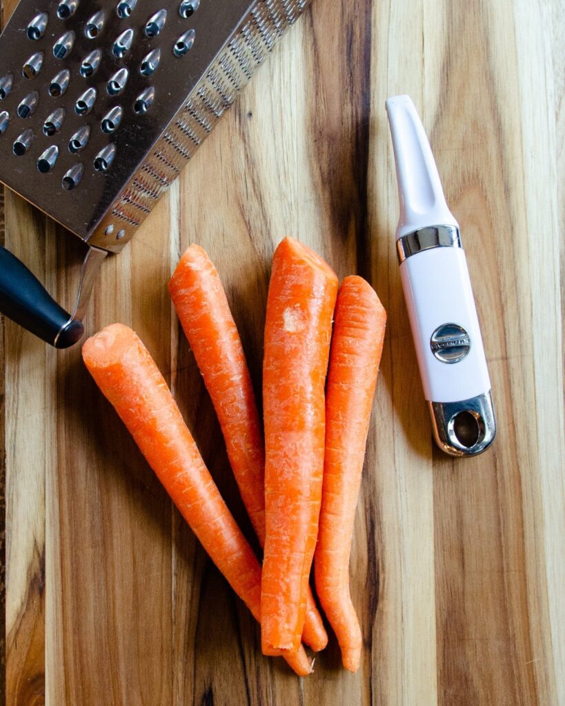 Carrots on a wooden cutting board with a peeler and a grater.