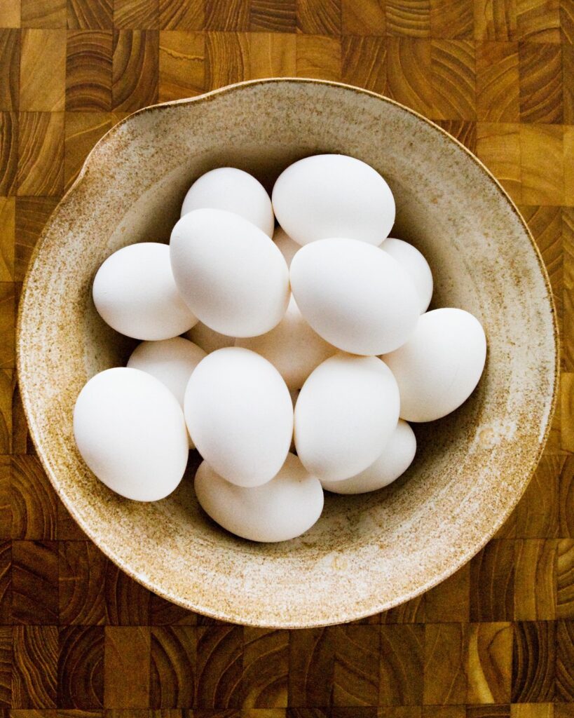 A ceramic bowl of white eggs on a wooden cutting board.