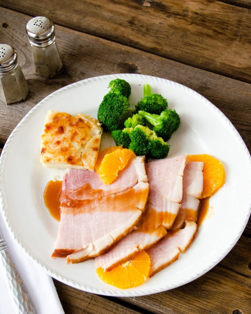 Sliced baked ham with madeira and oranges on a white plate with potato gratin and broccoli.